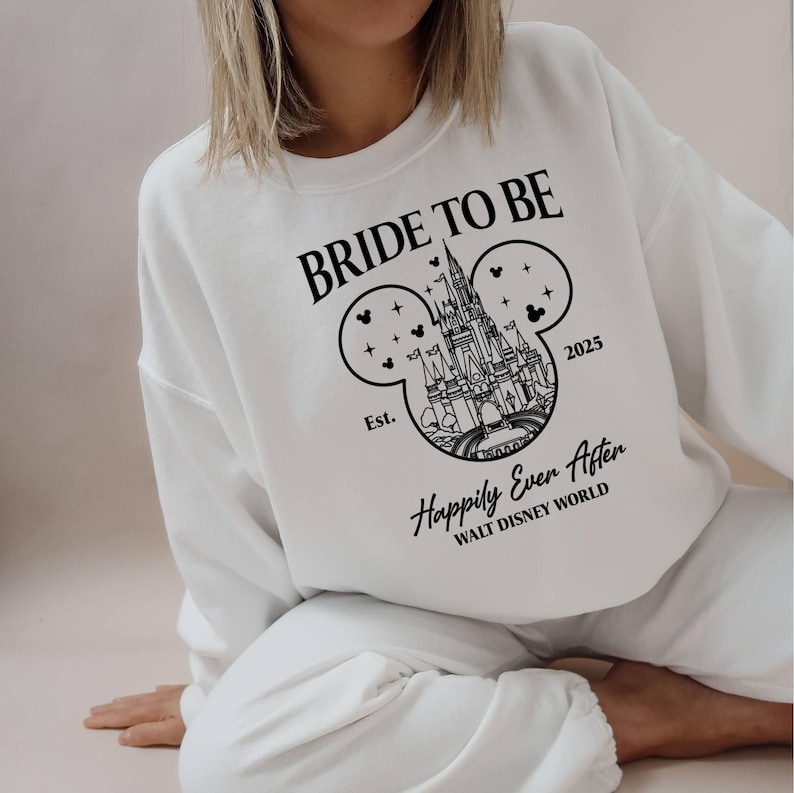 Puede incluir: Sudadera blanca con texto negro que dice "Bride to Be" y un contorno negro del castillo de Cenicienta dentro de una oreja de Mickey Mouse. El texto "Est. 2025 Happily Ever After Walt Disney World" tambi&eacute;n est&aacute; impreso en la sudadera.