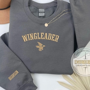 May include: Gray sweatshirt with gold embroidery that reads "WINGLEADER" with a dragon design. The sweatshirt has a gold embroidered "ROORSON" on the left sleeve.