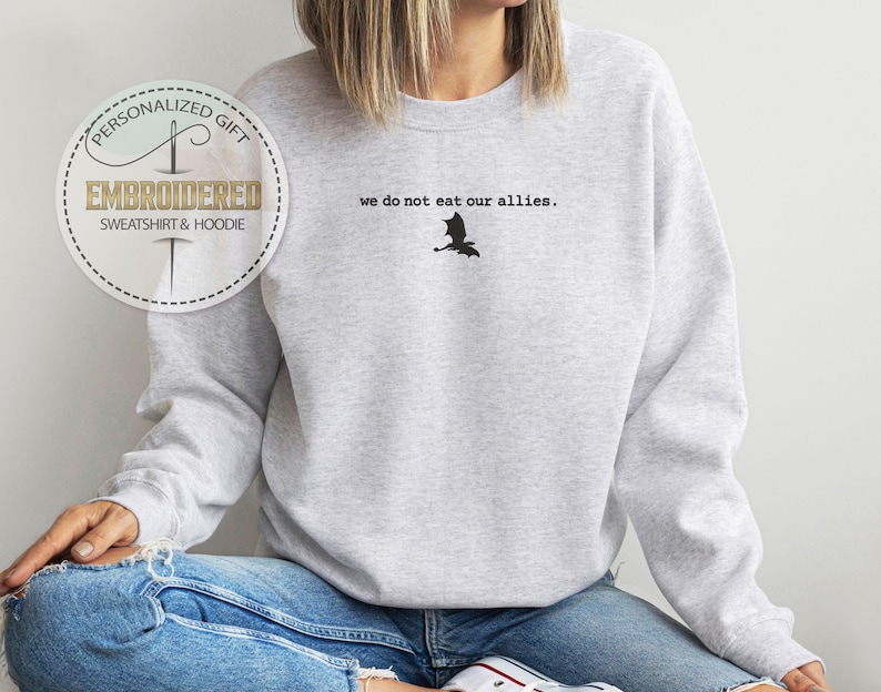 Puede incluir: Una sudadera gris con el texto "we do not eat our allies." y una silueta negra de un drag&oacute;n.