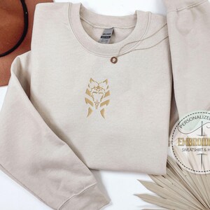 May include: A beige sweatshirt with a gold embroidered design of a Star Wars character's face. The text "Personalized Gift Embroidered Sweatshirt & Hoodie" is on a brown circle sticker.
