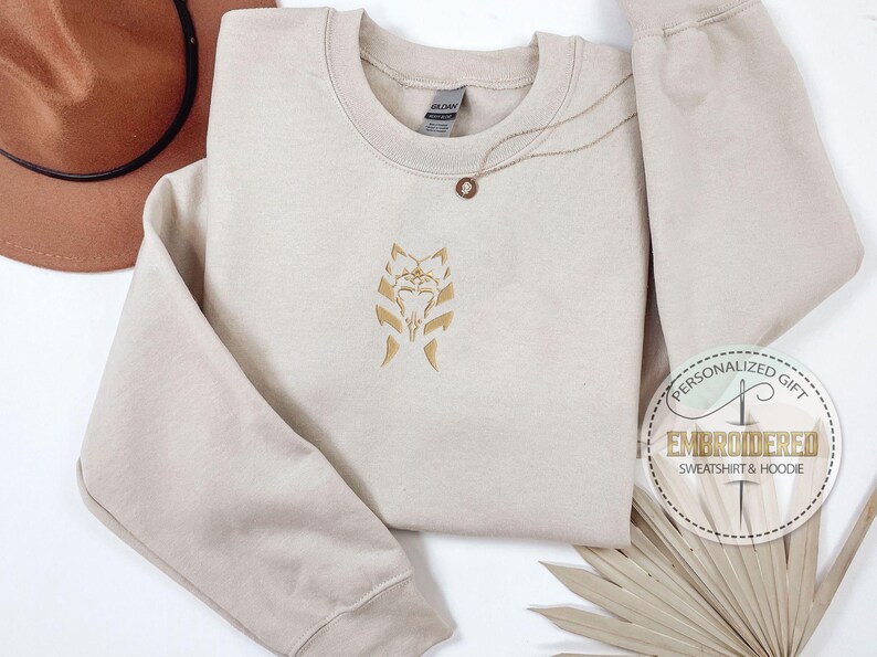 May include: A beige sweatshirt with a gold embroidered design of a Star Wars character's face. The text "Personalized Gift Embroidered Sweatshirt & Hoodie" is on a brown circle sticker.