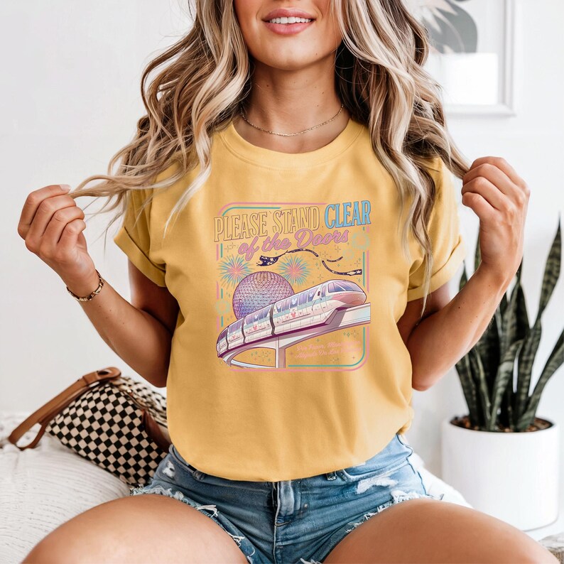 May include: Mustard yellow t-shirt with a graphic featuring a monorail, a sphere, and the text "PLEASE STAND CLEAR of the Doors". The design is framed in a rectangle with pastel colors.