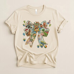 May include: A beige t-shirt featuring a colorful graphic of a bow filled with various Toy Story characters. The design includes Woody, Buzz Lightyear, and other characters, along with balloons and a rocket ship. The shirt has short sleeves and a crew neck.