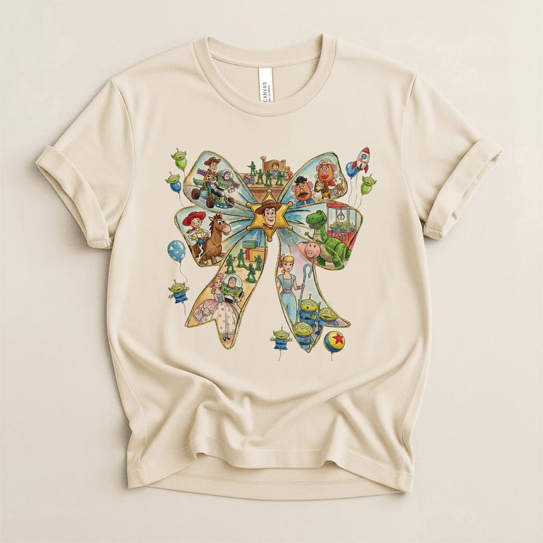 May include: A beige t-shirt featuring a colorful graphic of a bow filled with various Toy Story characters. The design includes Woody, Buzz Lightyear, and other characters, along with balloons and a rocket ship. The shirt has short sleeves and a crew neck.