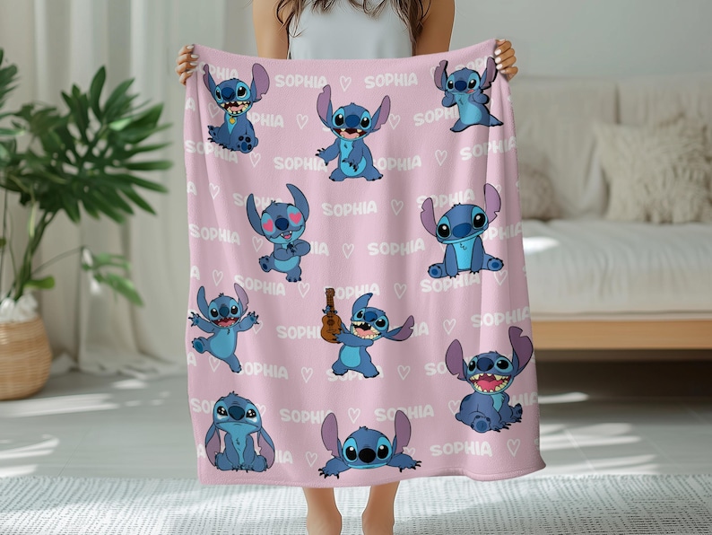May include: A pink blanket featuring multiple blue cartoon character illustrations and the name "SOPHIA" repeated with heart accents. The blanket is held up, showcasing the design.