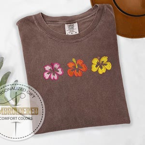 May include: Brown Comfort Colors t-shirt with embroidered hibiscus flowers in pink, orange, and yellow. The shirt has a crew neck and a tag that says "Comfort Colors". The shirt is a personalized gift.