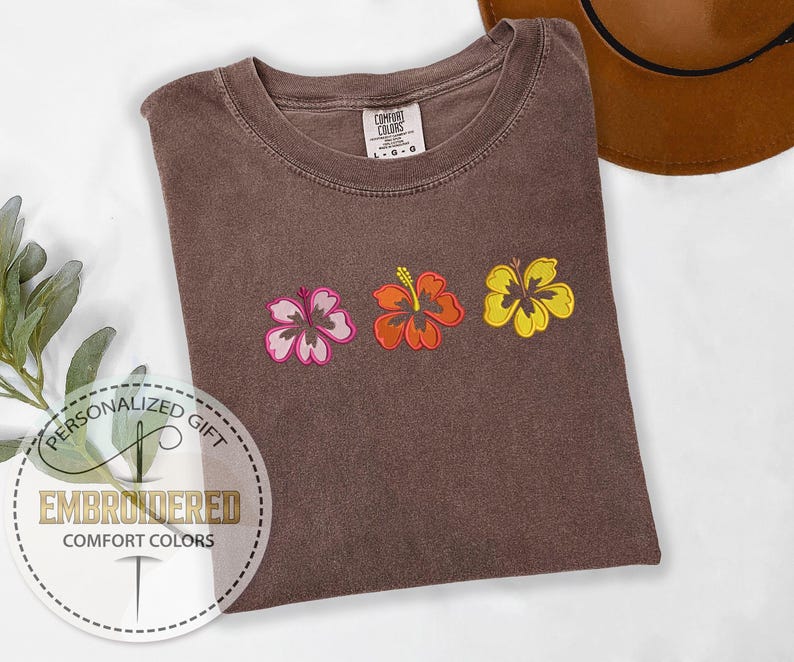 May include: Brown Comfort Colors t-shirt with embroidered hibiscus flowers in pink, orange, and yellow. The shirt has a crew neck and a tag that says "Comfort Colors". The shirt is a personalized gift.