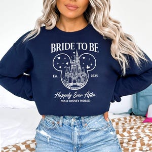 Puede incluir: Una sudadera azul marino con el texto "Bride to Be" y un contorno blanco del castillo de Cenicienta dentro de las orejas de Mickey Mouse. El texto "Est. 2025" y "Happily Ever After WALT DISNEY WORLD" tambi&eacute;n est&aacute;n impresos en la sudadera.