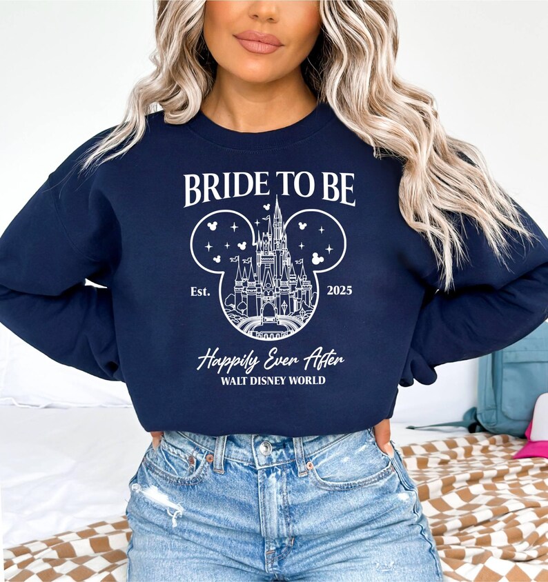 Puede incluir: Una sudadera azul marino con el texto "Bride to Be" y un contorno blanco del castillo de Cenicienta dentro de las orejas de Mickey Mouse. El texto "Est. 2025" y "Happily Ever After WALT DISNEY WORLD" tambi&eacute;n est&aacute;n impresos en la sudadera.