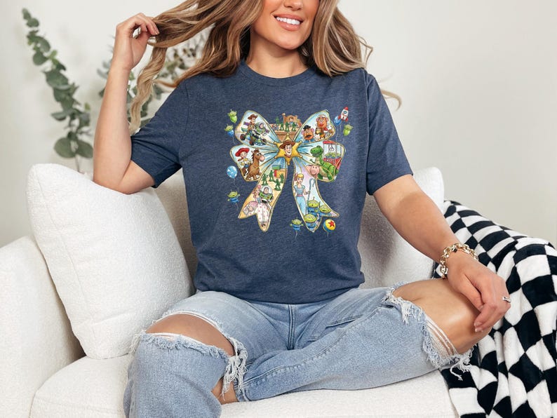 May include: A navy blue t-shirt featuring a colorful bow design filled with cartoon characters. The bow design includes characters from the Toy Story movie franchise. The shirt is made of a soft material.