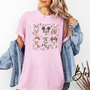 May include: Light pink t-shirt featuring a graphic of Disney characters including Mickey Mouse, Minnie Mouse, Donald Duck, and Pluto. The design includes hearts and signatures. The shirt is a casual, oversized fit.