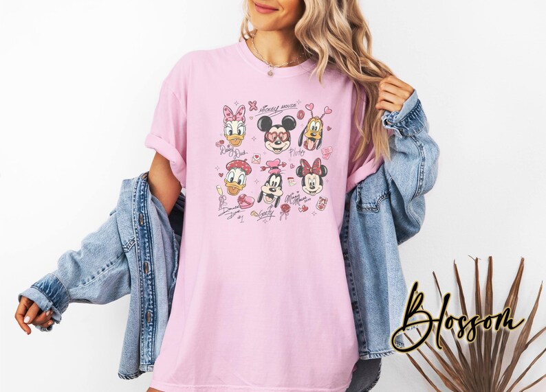 May include: Light pink t-shirt featuring a graphic of Disney characters including Mickey Mouse, Minnie Mouse, Donald Duck, and Pluto. The design includes hearts and signatures. The shirt is a casual, oversized fit.