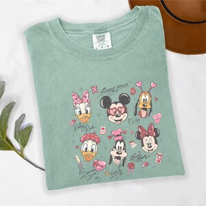 May include: A sage green t-shirt featuring a graphic of Disney characters, including Mickey Mouse, Minnie Mouse, Donald Duck, Daisy Duck, Pluto, and Goofy. The design incorporates hearts and the words "Mickey Mouse" and character names.