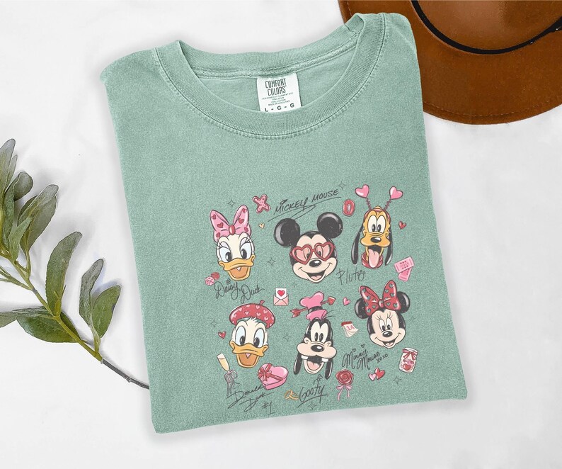 May include: A sage green t-shirt featuring a graphic of Disney characters, including Mickey Mouse, Minnie Mouse, Donald Duck, Daisy Duck, Pluto, and Goofy. The design incorporates hearts and the words "Mickey Mouse" and character names.