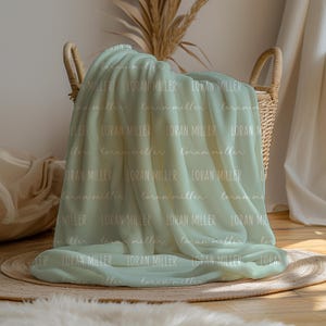 May include: A soft, light green blanket featuring a repeating pattern of the name LORAN MILLER printed in both a clean sans-serif font and a cursive script. The fabric is draped over a woven wicker basket, creating folds and soft shadows.