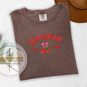 Pu&ograve; includere: Una maglietta marrone con il testo "Disneyland Est. 1955" ricamato in rosso. Il testo &egrave; arcuato e ha una ciliegia rossa con un fiocco rosa su ogni lato.