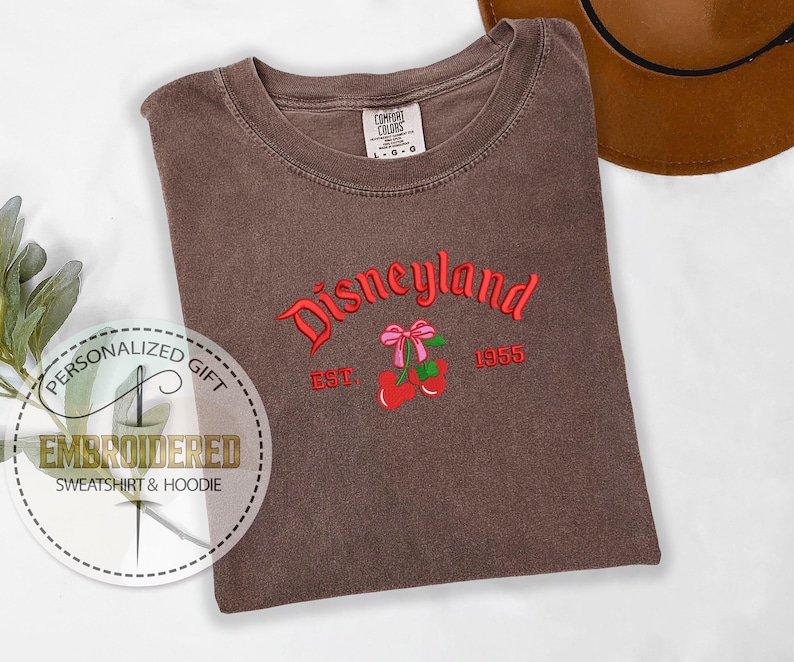 Pu&ograve; includere: Una maglietta marrone con il testo "Disneyland Est. 1955" ricamato in rosso. Il testo &egrave; arcuato e ha una ciliegia rossa con un fiocco rosa su ogni lato.