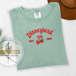 Pu&ograve; includere: Una maglietta verde chiaro con il testo "Disneyland" in lettere rosse e nere. Il testo "Est. 1955" &egrave; sotto il testo Disneyland. Una ciliegia rossa con un fiocco rosa &egrave; ricamata sulla maglietta.