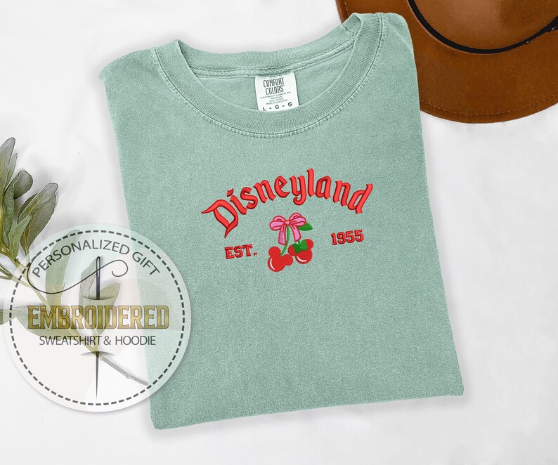 Pu&ograve; includere: Una maglietta verde chiaro con il testo "Disneyland" in lettere rosse e nere. Il testo "Est. 1955" &egrave; sotto il testo Disneyland. Una ciliegia rossa con un fiocco rosa &egrave; ricamata sulla maglietta.
