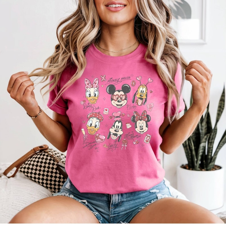 May include: A pink t-shirt featuring a collage of Disney characters, including Mickey Mouse, Minnie Mouse, Donald Duck, and Daisy Duck. The design includes hearts and the text "Mickey Mouse".