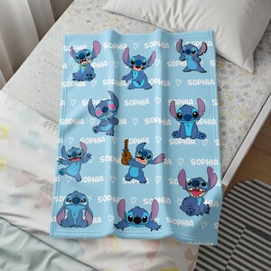 May include: A light blue blanket featuring multiple illustrations of the character Stitch in various poses. The blanket has the name "SOPHIA" and heart symbols printed across it. The blanket is made of a soft material.