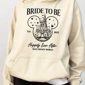 Puede incluir: Una sudadera con capucha beige con un gr&aacute;fico negro de las orejas de Mickey Mouse con el castillo de Cenicienta en su interior. El texto "Bride to Be" est&aacute; por encima del gr&aacute;fico y "Est. 2025" est&aacute; por debajo. El texto "Happily Ever After" est&aacute; debajo del gr&aacute;fico y "Walt Disney World" est&aacute; debajo.