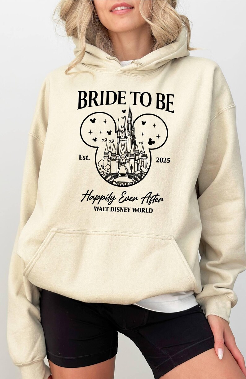 Puede incluir: Una sudadera con capucha beige con un gr&aacute;fico negro de las orejas de Mickey Mouse con el castillo de Cenicienta en su interior. El texto "Bride to Be" est&aacute; por encima del gr&aacute;fico y "Est. 2025" est&aacute; por debajo. El texto "Happily Ever After" est&aacute; debajo del gr&aacute;fico y "Walt Disney World" est&aacute; debajo.