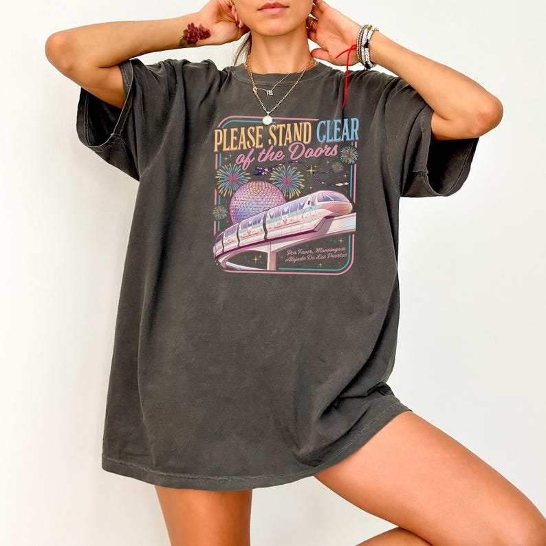 May include: Dark grey t-shirt with a colorful graphic featuring a monorail, fireworks, and the text "PLEASE STAND CLEAR of the DOORS". The design includes a stylized depiction of a globe and text at the bottom.