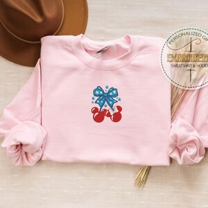 Op de afbeelding: Een lichtroze sweatshirt met een ronde hals en lange mouwen. De voorkant heeft een geborduurd ontwerp van een blauwe strik met witte sterren en rode Mickey Mouse-achtige vormen. Een bruine hoed staat op de achtergrond.