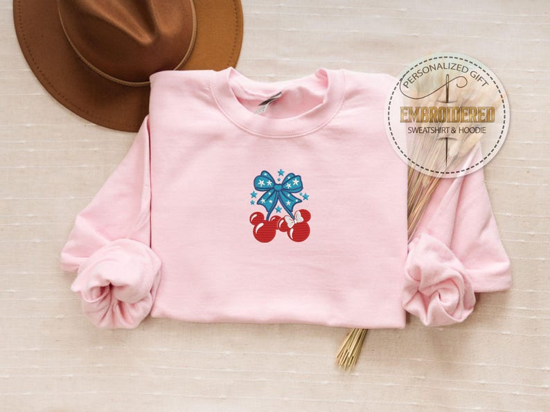 Op de afbeelding: Een lichtroze sweatshirt met een ronde hals en lange mouwen. De voorkant heeft een geborduurd ontwerp van een blauwe strik met witte sterren en rode Mickey Mouse-achtige vormen. Een bruine hoed staat op de achtergrond.