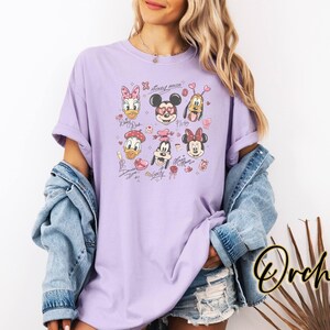 May include: A light purple t-shirt featuring cartoon character faces, including Mickey and Minnie Mouse, Donald Duck, and Pluto. The design includes hearts and signatures. The word "Orchid" is written in a cursive font on the bottom right.