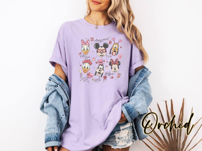 May include: A light purple t-shirt featuring cartoon character faces, including Mickey and Minnie Mouse, Donald Duck, and Pluto. The design includes hearts and signatures. The word "Orchid" is written in a cursive font on the bottom right.
