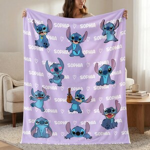 May include: A lavender throw blanket featuring multiple images of a blue cartoon character. The blanket has the name "SOPHIA" and white hearts printed across the surface. The blanket is made of a soft material.