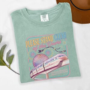 May include: A light green t-shirt with the text "PLEASE STAND CLEAR of the DOORS" above a graphic of a monorail and a large sphere. The shirt is a Comfort Colors brand, size L-G.