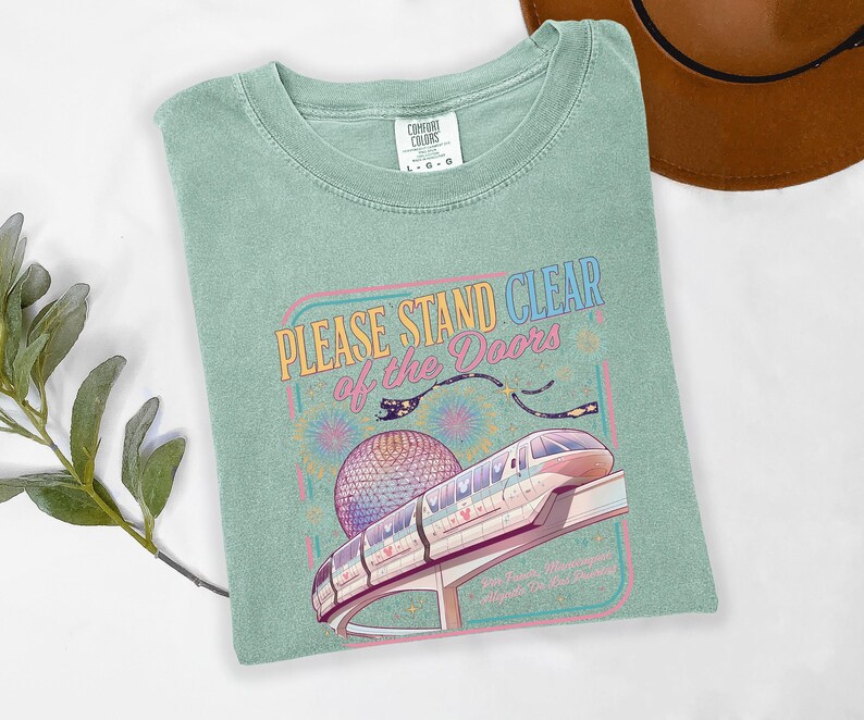 May include: A light green t-shirt with the text "PLEASE STAND CLEAR of the DOORS" above a graphic of a monorail and a large sphere. The shirt is a Comfort Colors brand, size L-G.