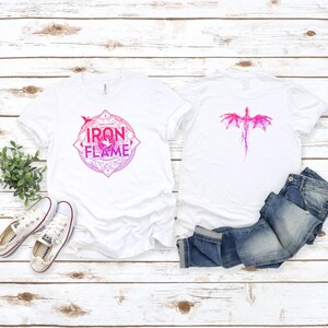 May include: Two white t-shirts with a pink and white dragon design. One shirt has the words "Iron & Flame" in a circle with a dragon inside. The shirts are laid out on a white wooden surface with a pair of white sneakers and blue jeans.