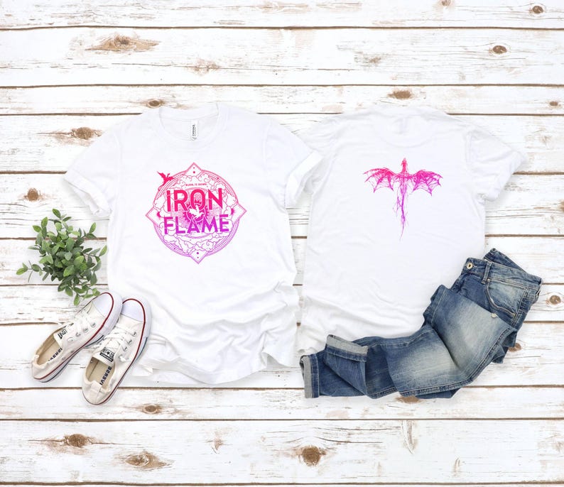May include: Two white t-shirts with a pink and white dragon design. One shirt has the words "Iron & Flame" in a circle with a dragon inside. The shirts are laid out on a white wooden surface with a pair of white sneakers and blue jeans.