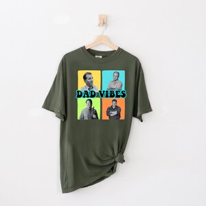 May include: Olive green t-shirt with a graphic design. The design features four black and white images of men in colorful squares, with the words "DAD VIBES" in teal and white text.