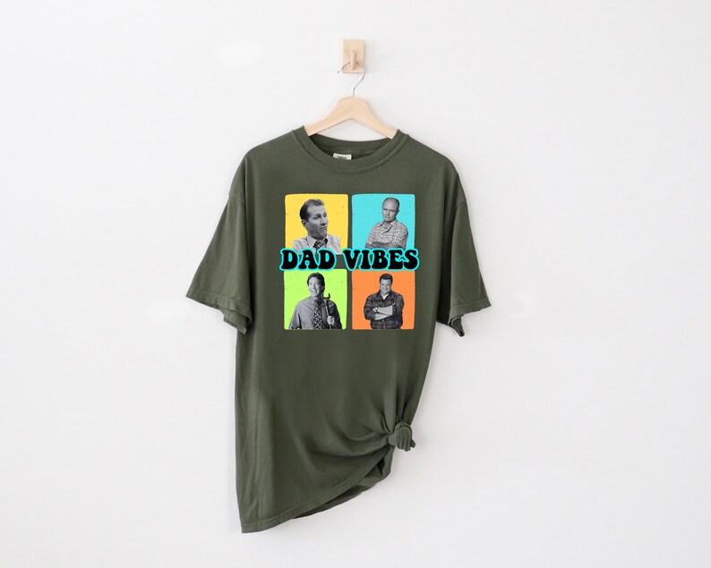 May include: Olive green t-shirt with a graphic design. The design features four black and white images of men in colorful squares, with the words "DAD VIBES" in teal and white text.