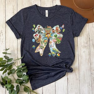 May include: Dark blue t-shirt featuring a colorful graphic of a bow filled with characters from the animated movie Toy Story. The graphic includes Woody, Buzz Lightyear, and other characters. The shirt is made of a soft material.