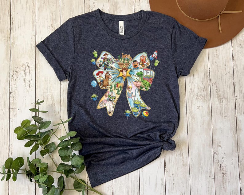May include: Dark blue t-shirt featuring a colorful graphic of a bow filled with characters from the animated movie Toy Story. The graphic includes Woody, Buzz Lightyear, and other characters. The shirt is made of a soft material.