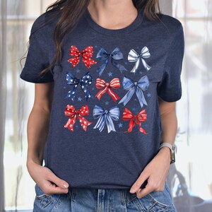 May include: Navy blue t-shirt with a pattern of red, white, and blue watercolour bows. The bows are arranged in a grid, with stars scattered around them. The shirt has a crew neck and short sleeves.