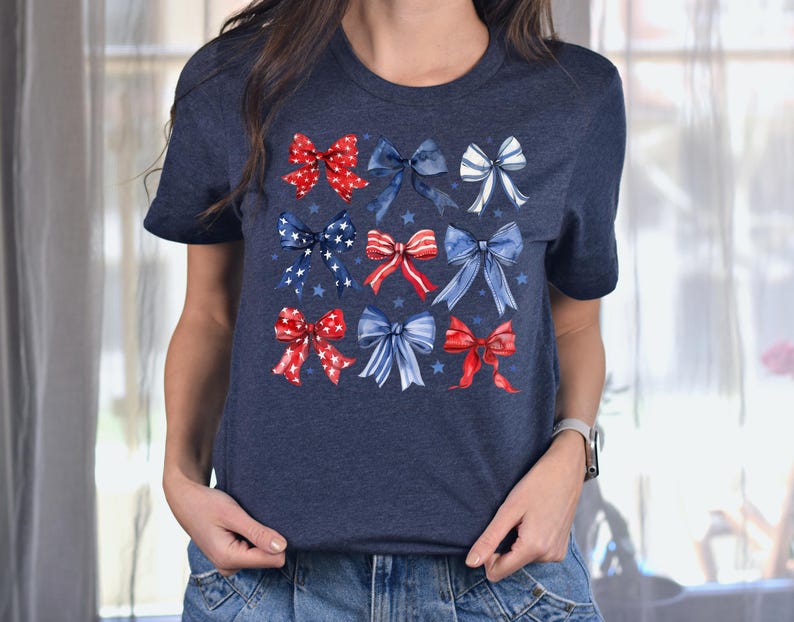 May include: Navy blue t-shirt with a pattern of red, white, and blue watercolour bows. The bows are arranged in a grid, with stars scattered around them. The shirt has a crew neck and short sleeves.