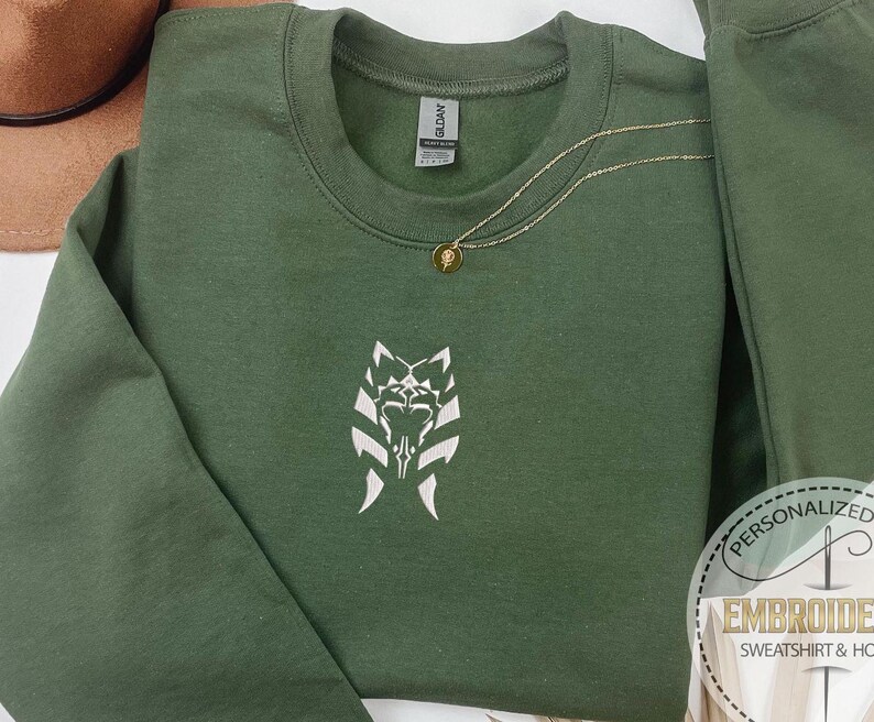 May include: A dark green sweatshirt with a white embroidered design of a stylized creature with a symbol on its chest. The sweatshirt has a gold chain necklace with a small gold pendant.