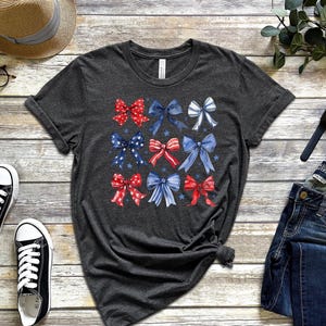 May include: Dark grey t-shirt with a grid of watercolour bows in red, white, and blue. The bows have star and stripe patterns. The shirt is displayed with black trainers, a straw hat, glasses, and a pair of blue jeans.