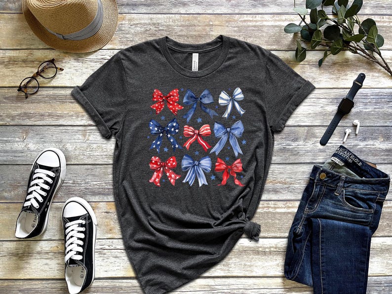 May include: Dark grey t-shirt with a grid of watercolour bows in red, white, and blue. The bows have star and stripe patterns. The shirt is displayed with black trainers, a straw hat, glasses, and a pair of blue jeans.
