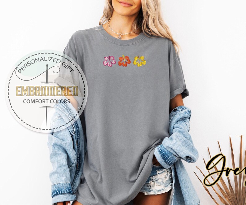 May include: A gray t-shirt with embroidered hibiscus flowers in pink, orange, and yellow. The shirt has the text "EMBROIDERED COMFORT COLORS" in a circular design. The shirt is part of the "Personalized Gift" collection.