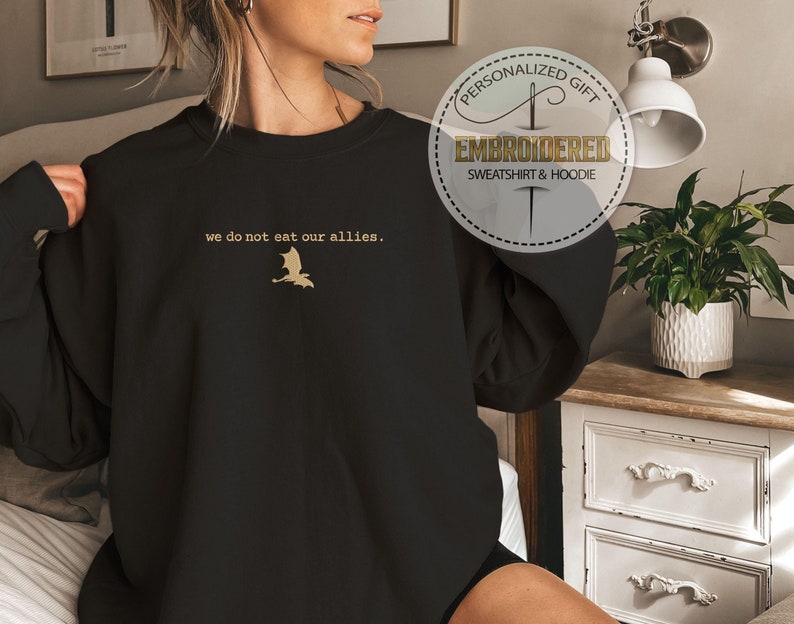 Puede incluir: Sudadera negra con un drag&oacute;n bordado en oro y el texto "we do not eat our allies."
