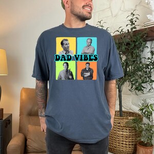 May include: Dark blue t-shirt with a graphic featuring four images of men and the text "DAD VIBES" in a teal font. The shirt is made of a soft material and has short sleeves. The shirt is a comfortable fit.