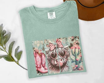 May include: A pale green Comfort Colors t-shirt with a graphic of a Highland cow wearing heart-shaped sunglasses and a pink bow. The graphic also includes a pink cowboy boot, cactus, and hearts.